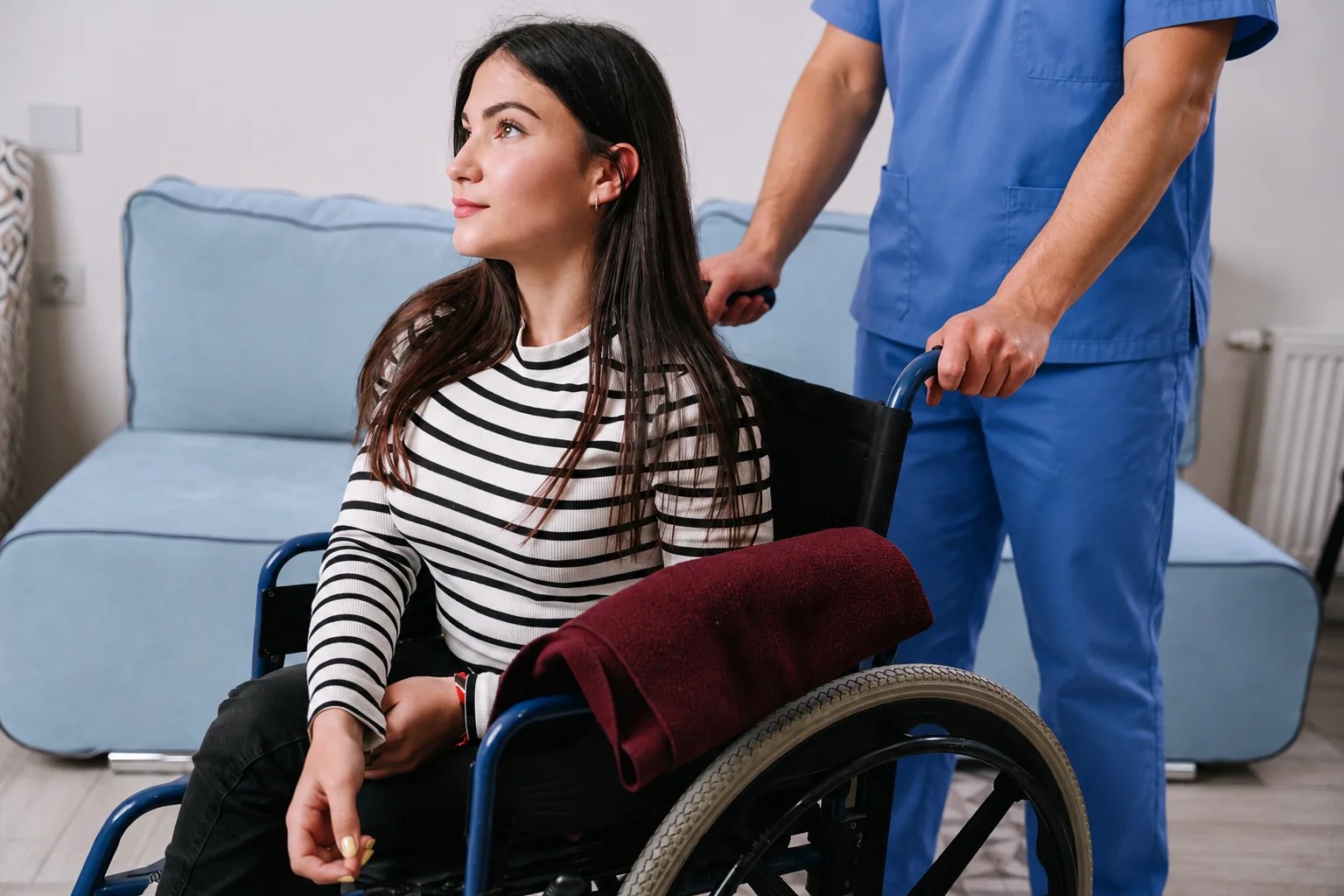 Care professional in blue scrubs assisting a woman using a wheelchair in a welcoming living room